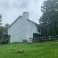A white two-story house with a brick chimney stands on a grassy hill next to a wooden fence and surrounded by trees.