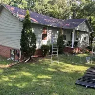 A light-gray, brick-bottomed house with a red roof stands behind a lawn, with a tall thin tree and a ladder in the yard.