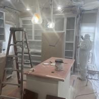 A worker in a protective suit sprays white paint onto kitchen cabinets in a room covered in plastic sheeting.