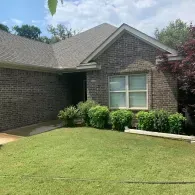 A single-story brick house with a gray shingled roof, a front lawn, and green bushes along the foundation.