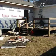 A house exterior under construction with a wooden deck, a ladder leaning against the wall, and debris scattered on the lawn.
