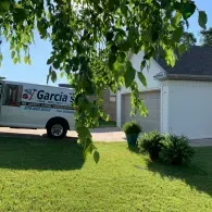 A Garcia service van parked in a driveway in front of a white house with a garage, framed by overhanging tree leaves.