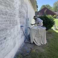A worker in a white hat uses a sprayer to apply white paint to a brick exterior wall next to a covered air conditioner.