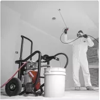 A worker in white protective gear uses an airless paint sprayer to apply finish to a white interior ceiling.
