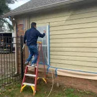 A person stands on a red stepladder, painting the exterior wall of a house with a spray gun near a masked-off window.