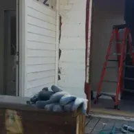 A pile of grey work gloves sits on a wooden ledge near a white exterior wall and a red stepladder.