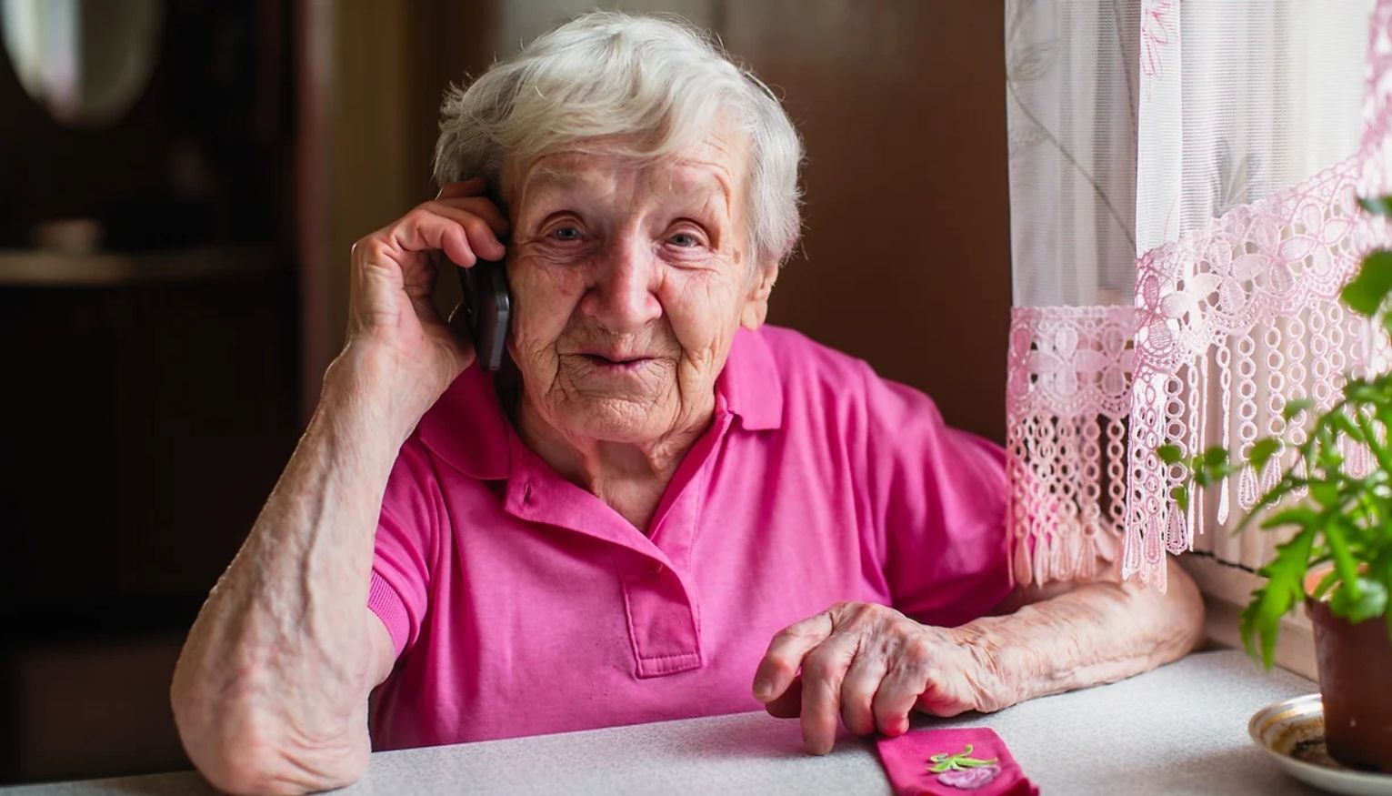 An elderly woman is sitting at a table talking on a cell phone.