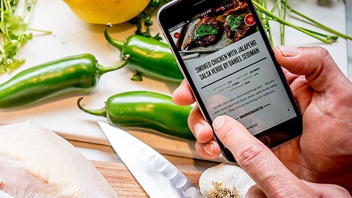 Image of a recipe on a smart phone in front of a cutting board with green chilies. 