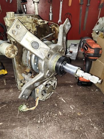 Mecanismo de accionamiento de lavadora desmontado. Componentes metálicos, cables eléctricos y herramientas en un banco de trabajo.