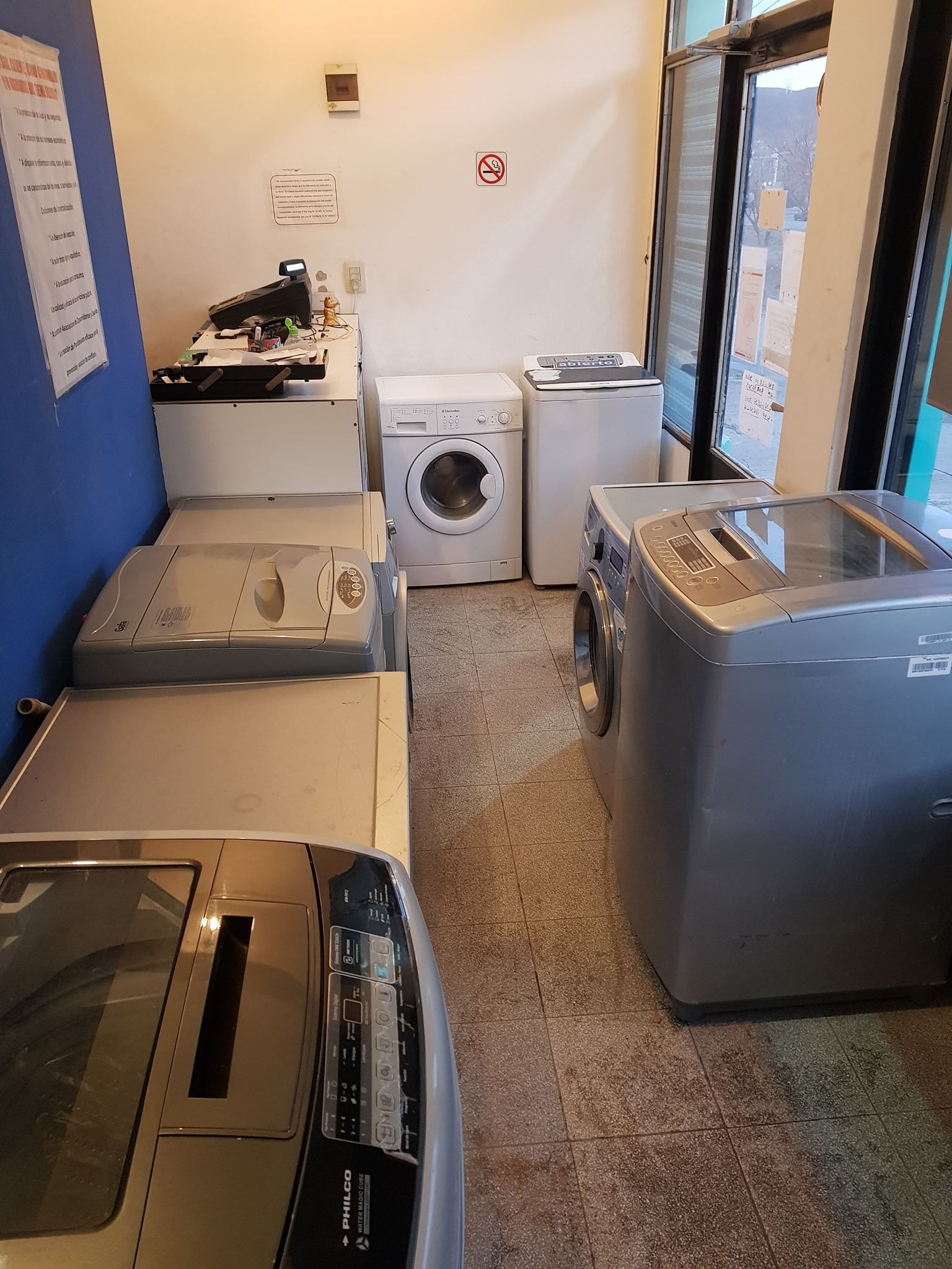 Interior de una lavandería con varias lavadoras en fila. Algunas son plateadas y otras blancas.