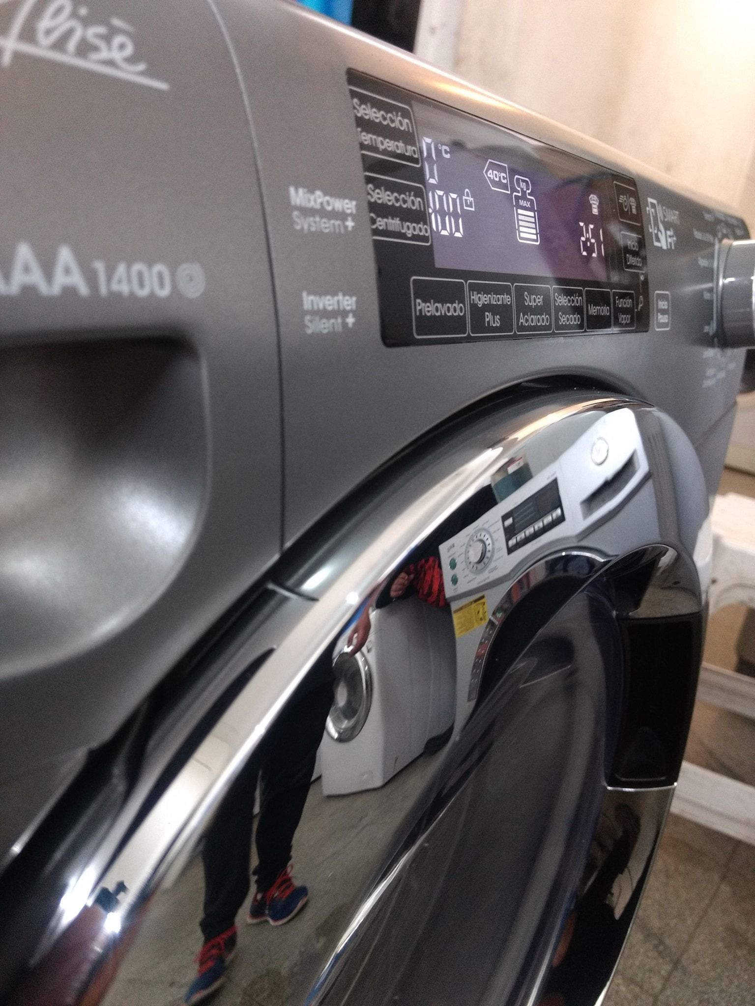 Primer plano de una lavadora plateada con pantalla digital, manija y una persona reflejada en el cromo.