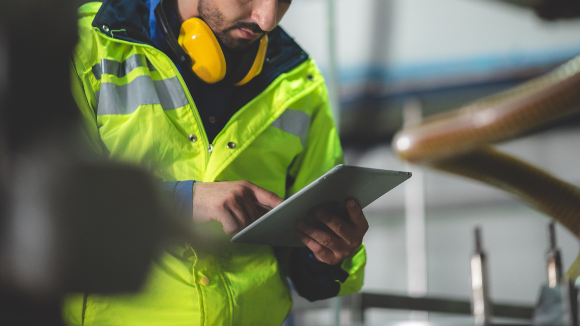 A man in a yellow jacket is using a tablet in a factory.