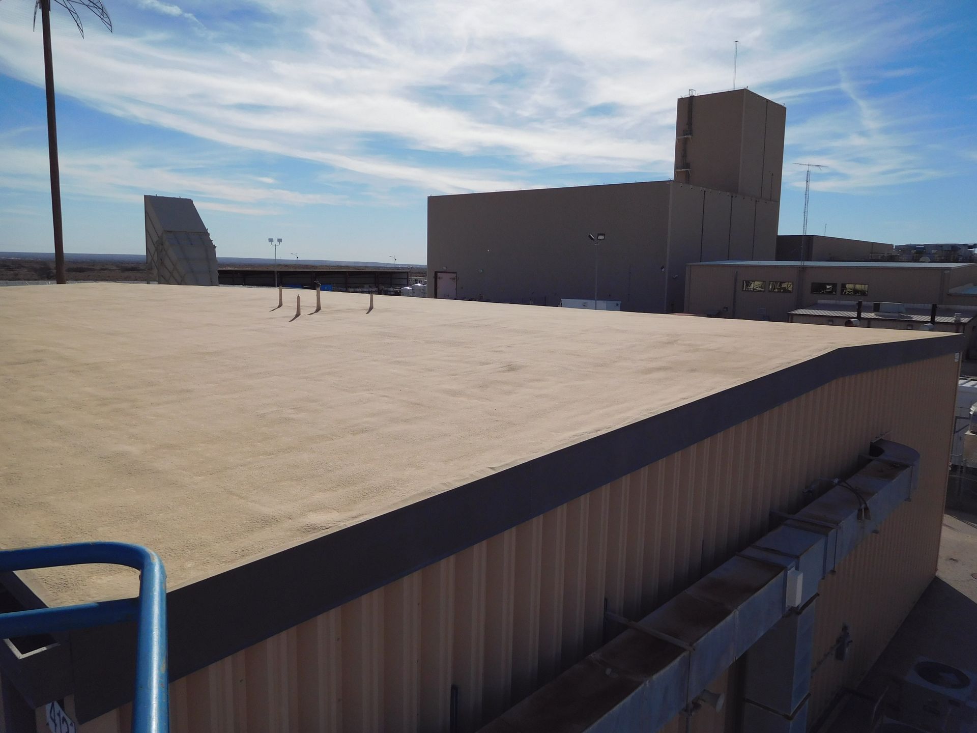 A flat tan roof with a brown edge. A building with a tower rises in the background under a blue sky.