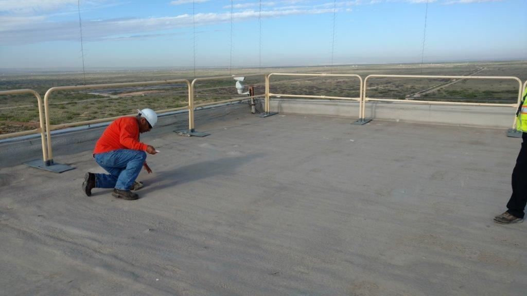 Man in red shirt kneels on a rooftop, holding a tool; another person stands nearby, background view of flat landscape.