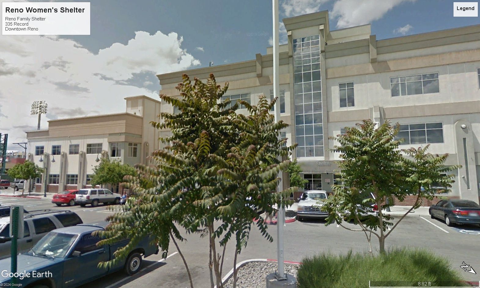 Building exterior with a light-colored facade, trees, parked cars, and a flagpole.