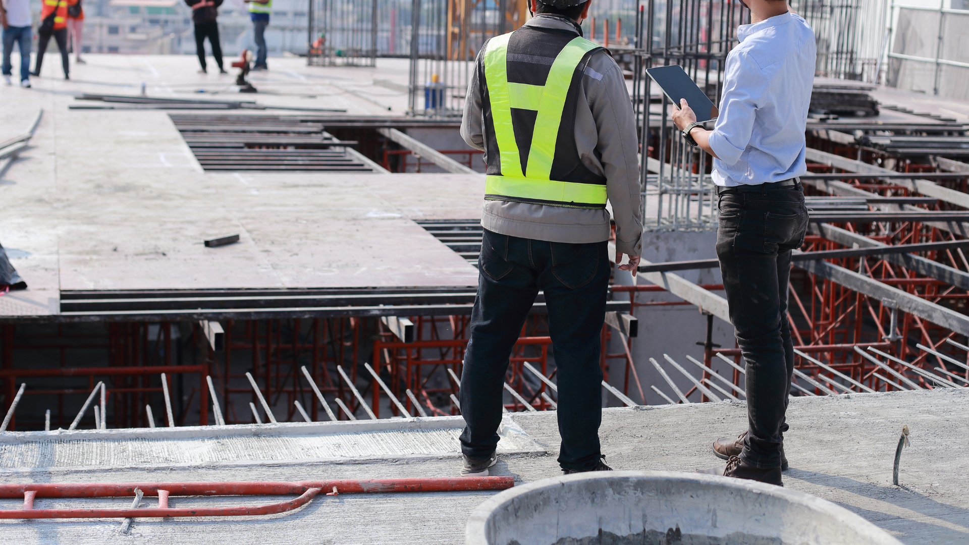 Two construction workers are standing on a construction site looking at a tablet.