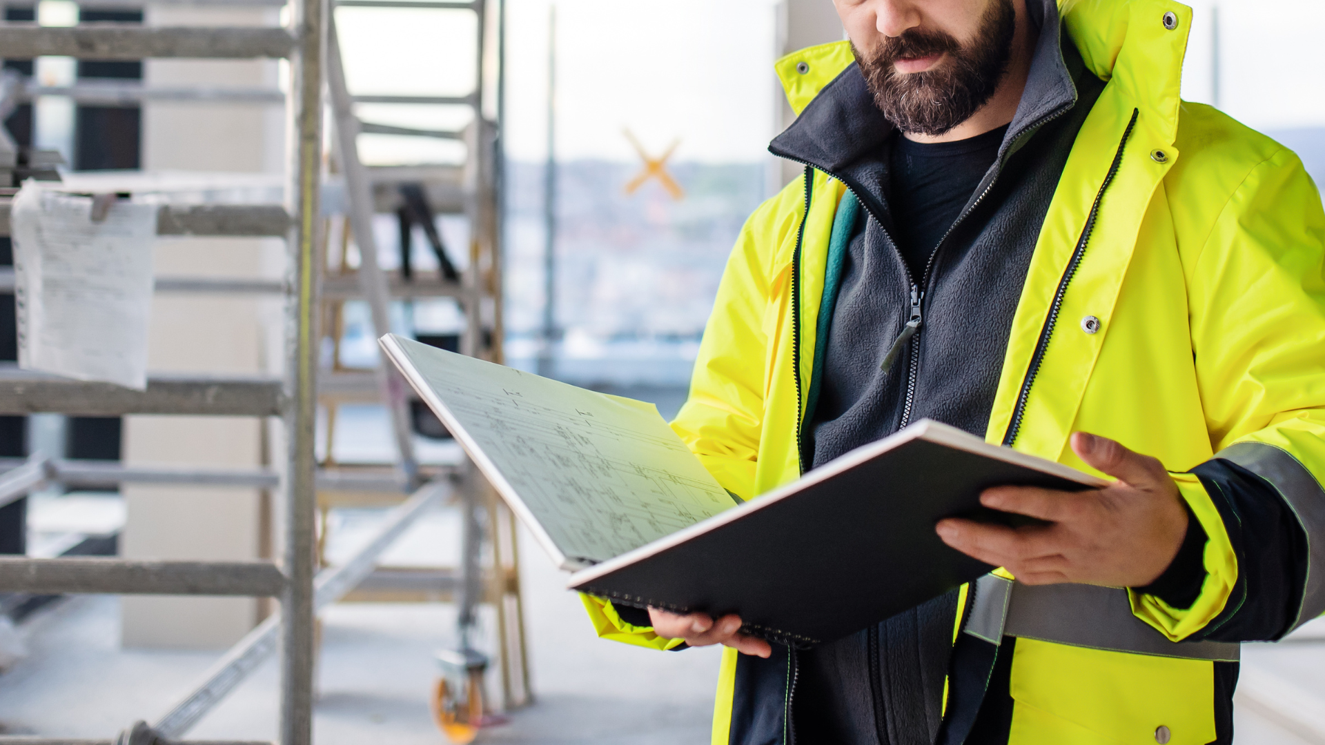A man in a yellow jacket is holding a clipboard and looking at it.