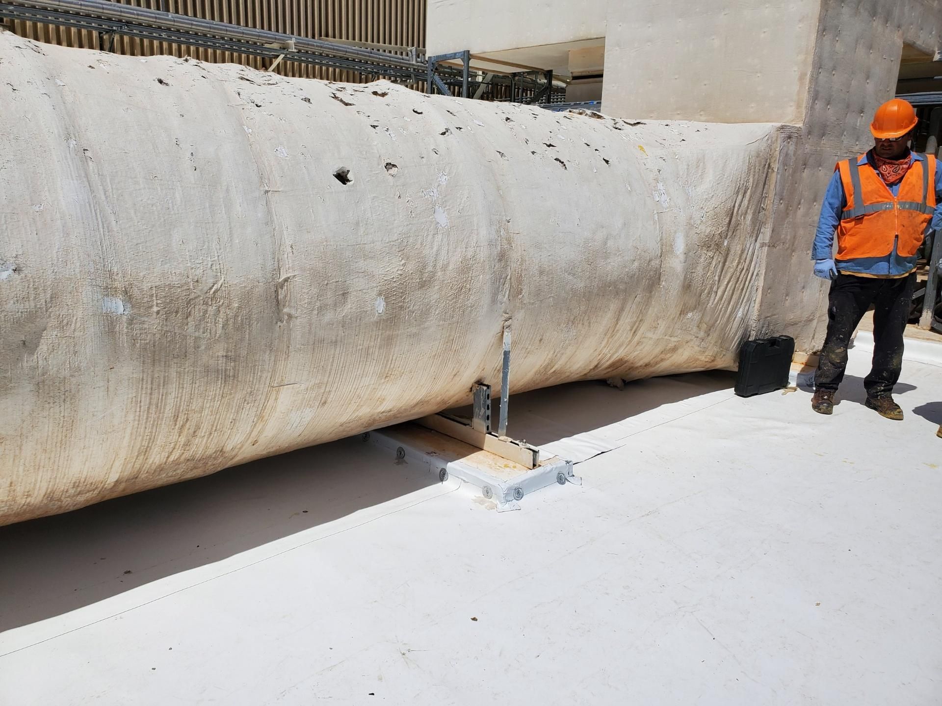 Construction worker in orange vest near large, sand-colored cylindrical structure on a white surface.