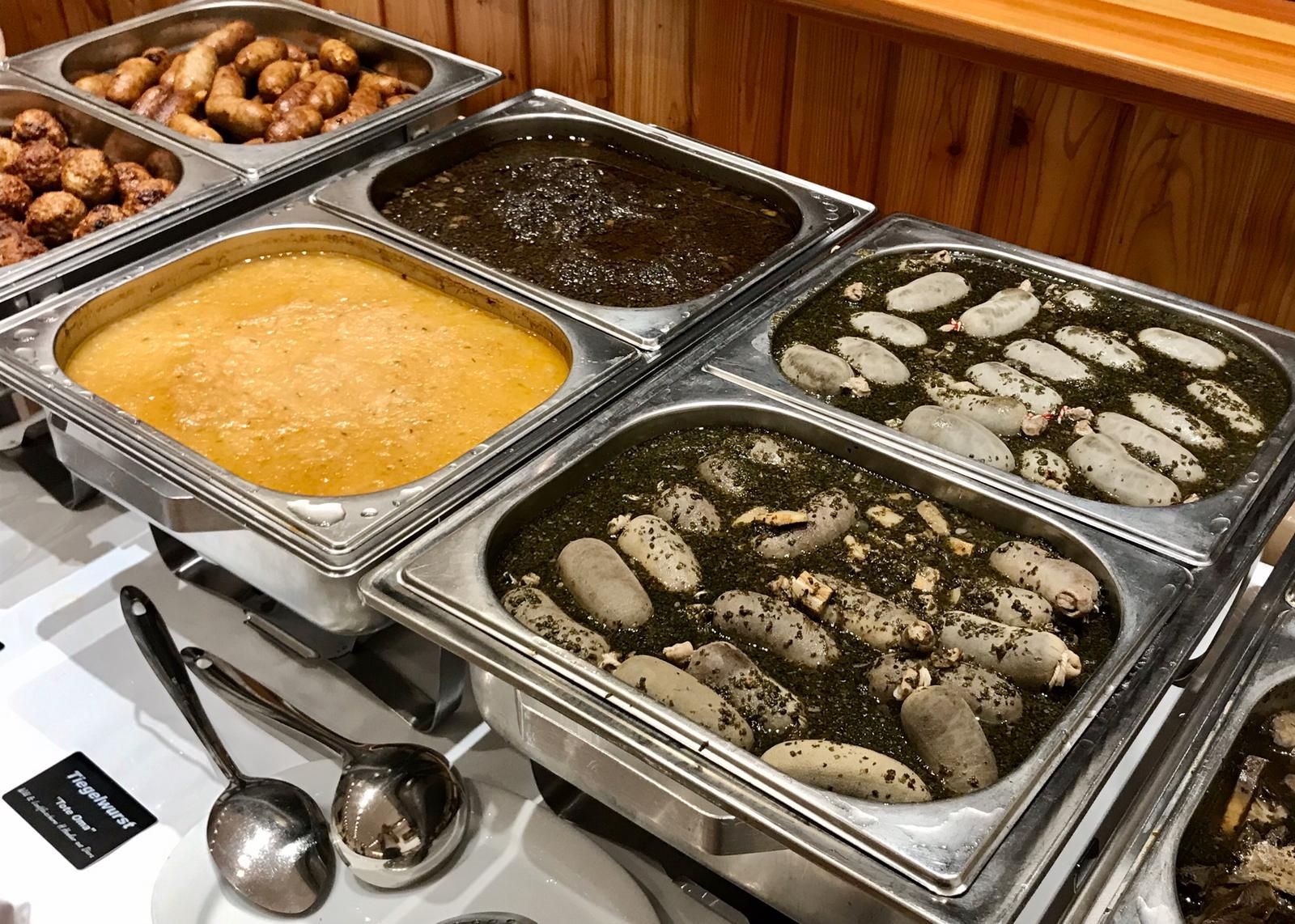Buffet mit spanischen Gerichten in Metallbehältern, darunter Suppen und Eintöpfe in verschiedenen Farben.