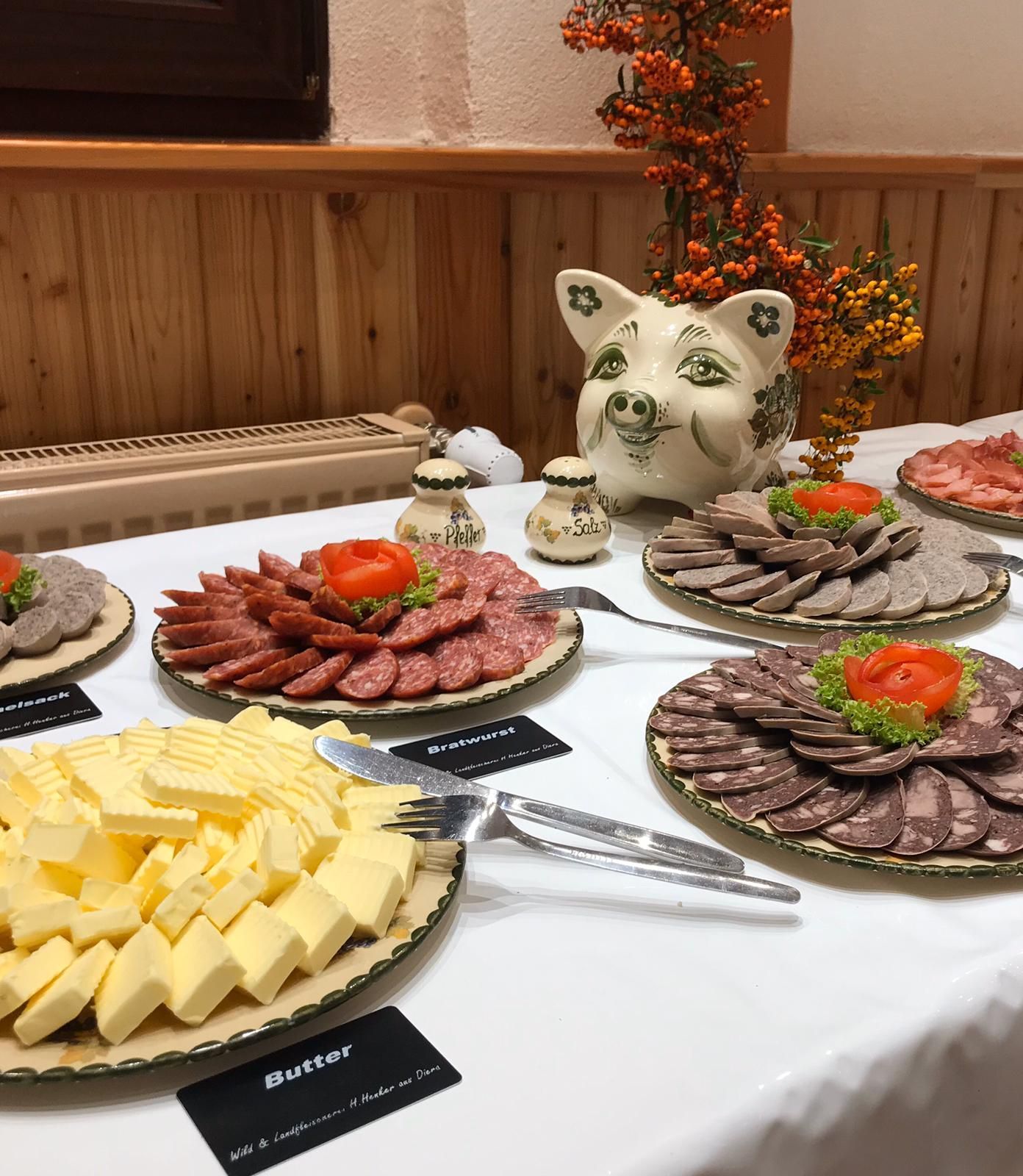 Ein Buffet mit Tellern mit Wurstscheiben, Butter und einem Sparschwein. Weiße Tischdecke, Holzwände und Herbstlaub im Hintergrund.