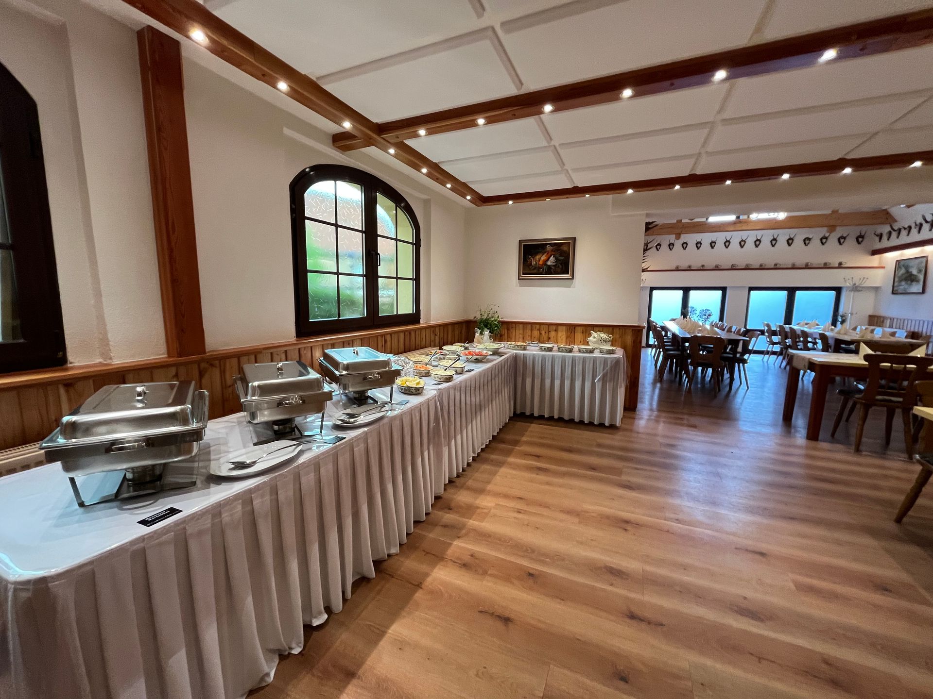 Buffettisch in einem Speisesaal mit Holzboden, mit Chafing Dishes und Speisen. Im Hintergrund sind Tische und Stühle aufgestellt.