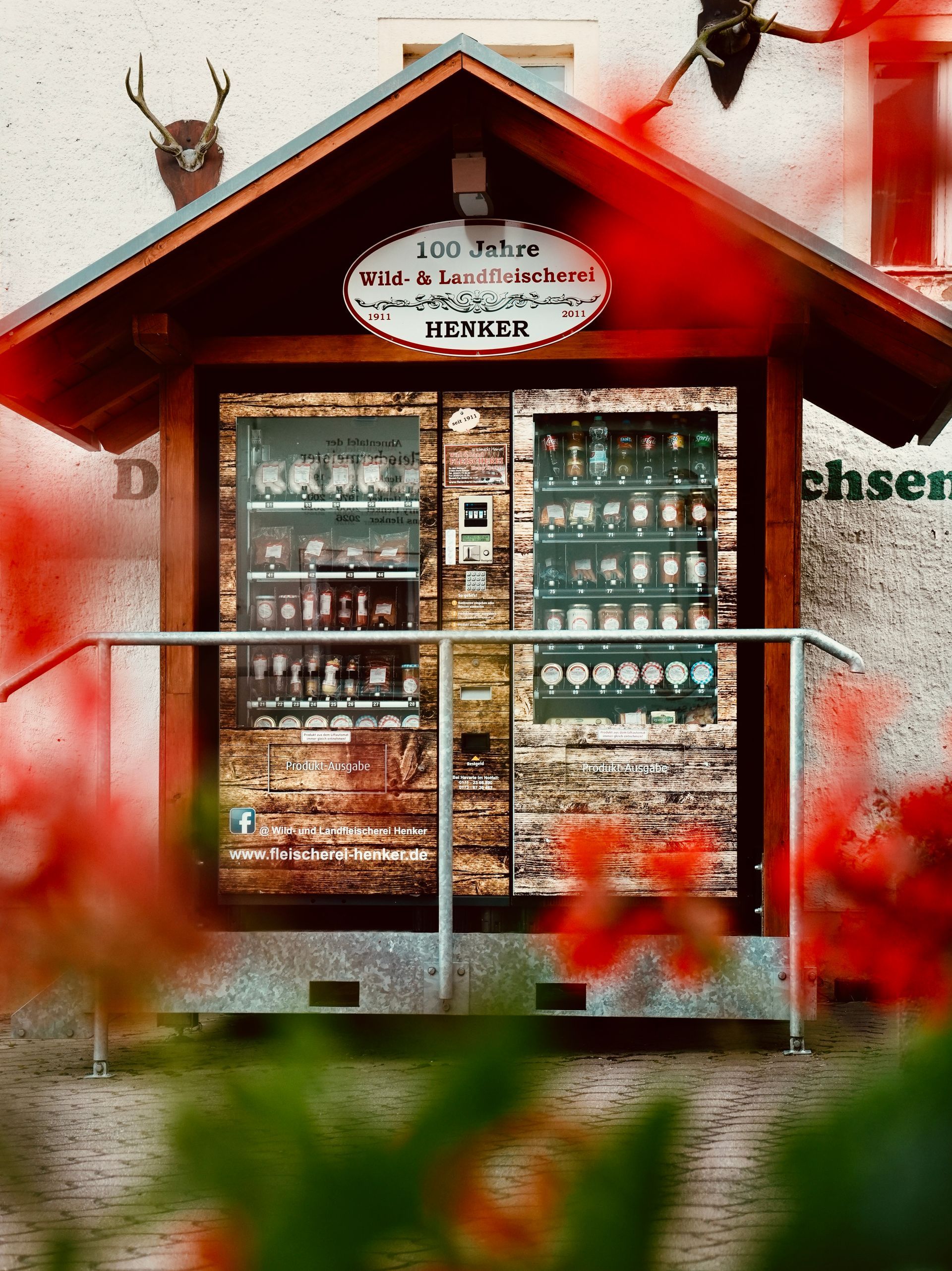 Hölzerner Getränkeautomat mit verschiedenen Getränken und Snacks, untergebracht in einem rustikalen Holzunterstand; im Vordergrund rote Blumen.