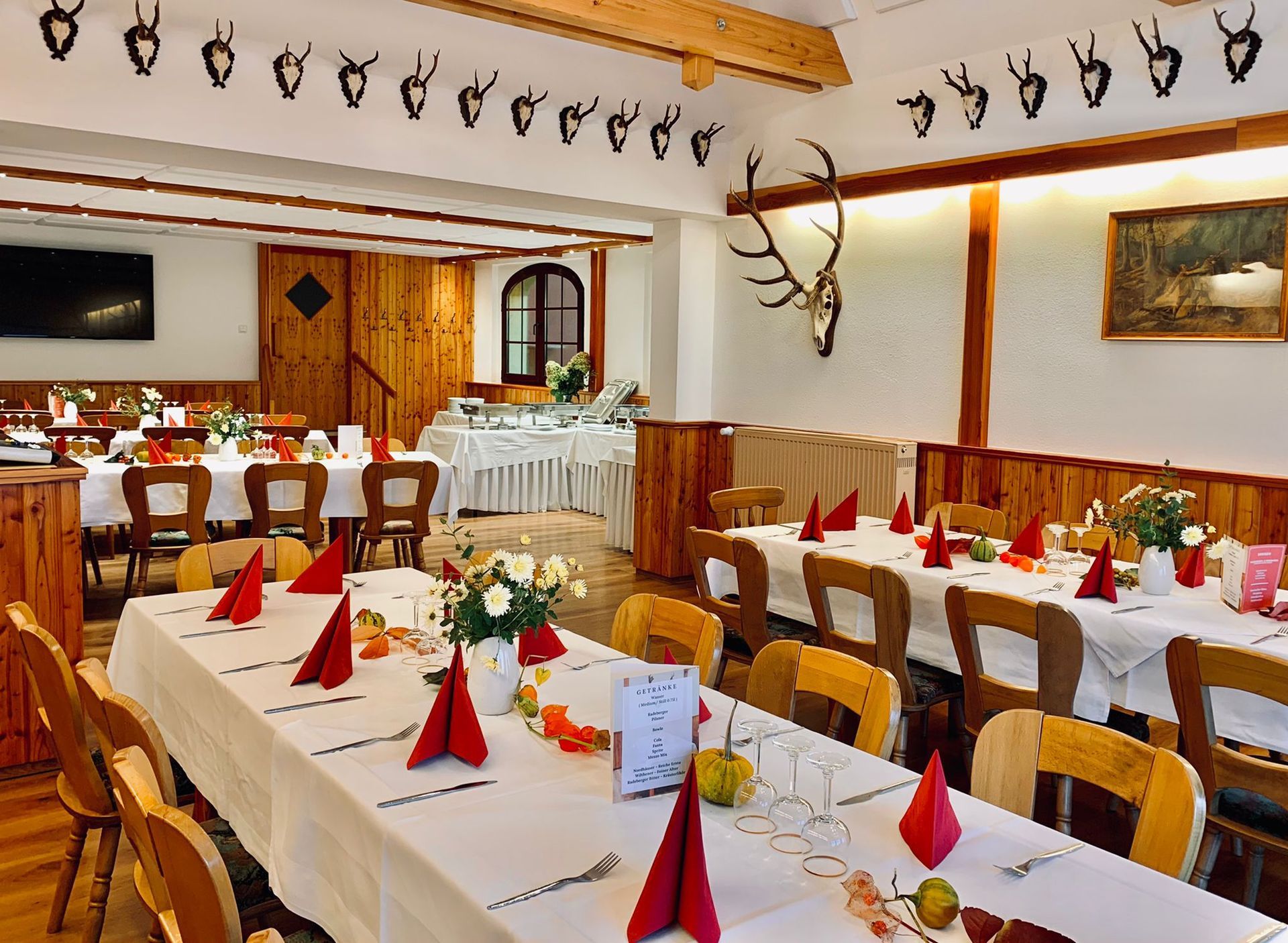 Restaurant-Innenraum mit langen Tischen, die für eine gesellige Runde gedeckt sind. Holzvertäfelungen, Geweihe und ein Buffet sorgen für eine rustikale Atmosphäre.