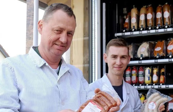 Zwei Männer in Metzgerkitteln, einer älter und einer jünger, stehen neben einem Fleischautomaten, halten Wurstpackungen in den Händen und lächeln.