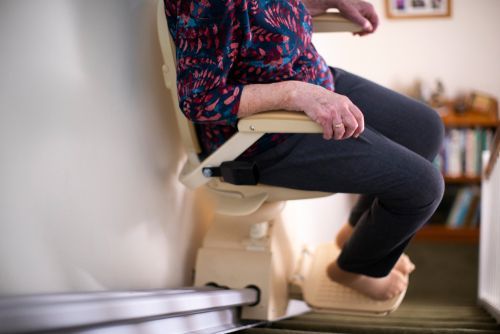 Person seated on a beige stair lift beside a staircase, wearing dark pants and a patterned top.