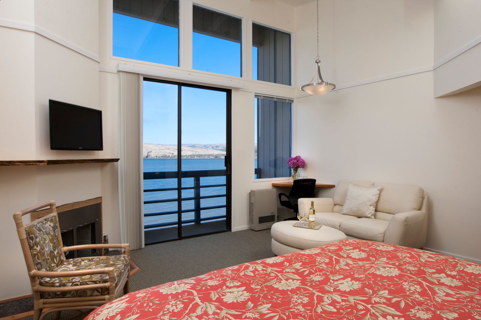 Bright hotel room with water view, sliding door, fireplace, couch, desk, and bed.
