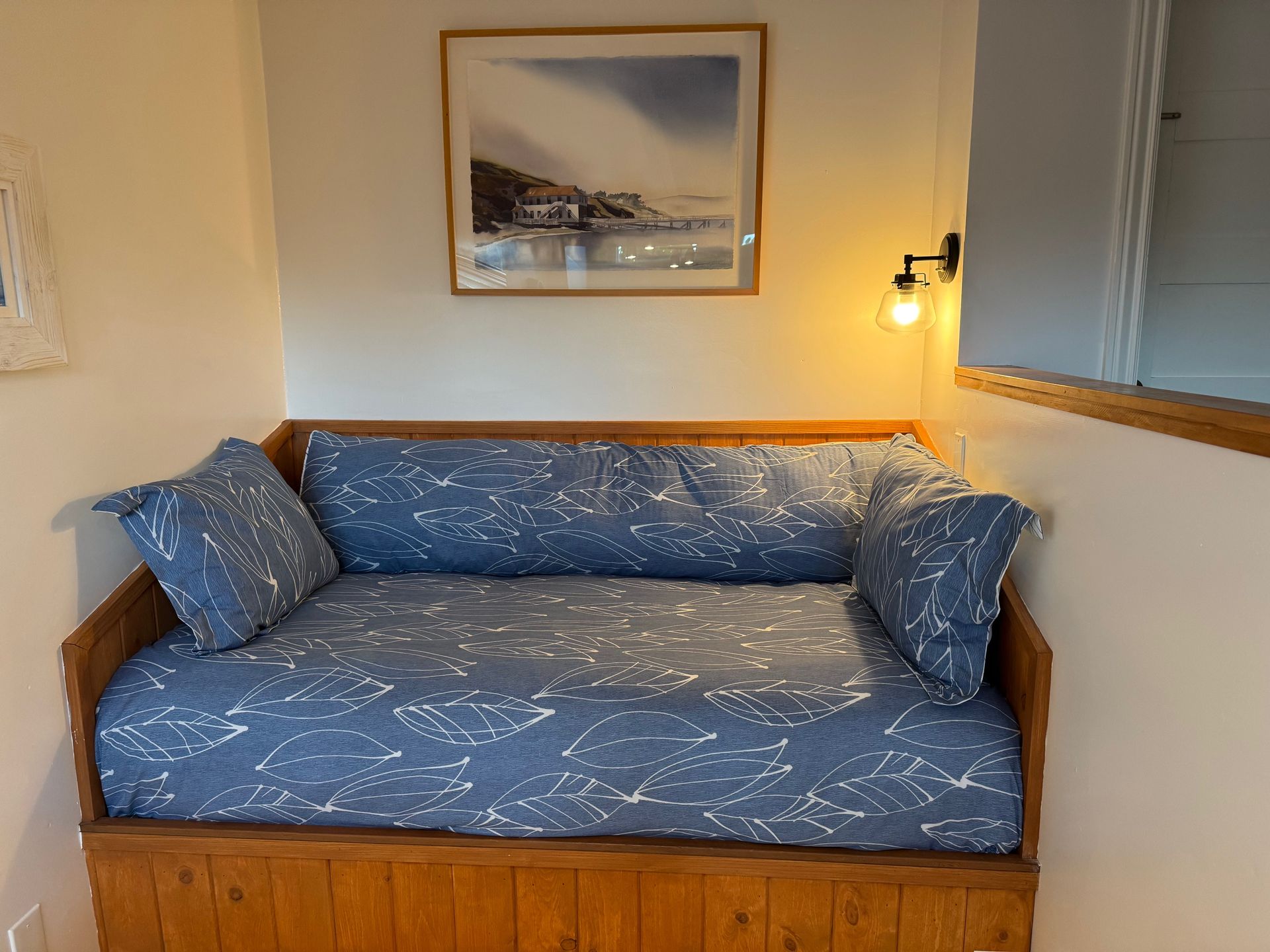 Wooden daybed with blue patterned cushions and pillows, beneath a watercolor landscape print.