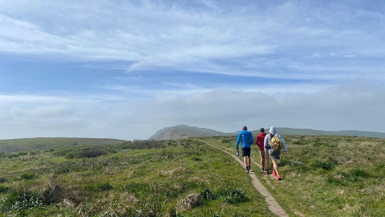 Three people walking on a grassy path toward a distant, rocky hill under a blue sky.