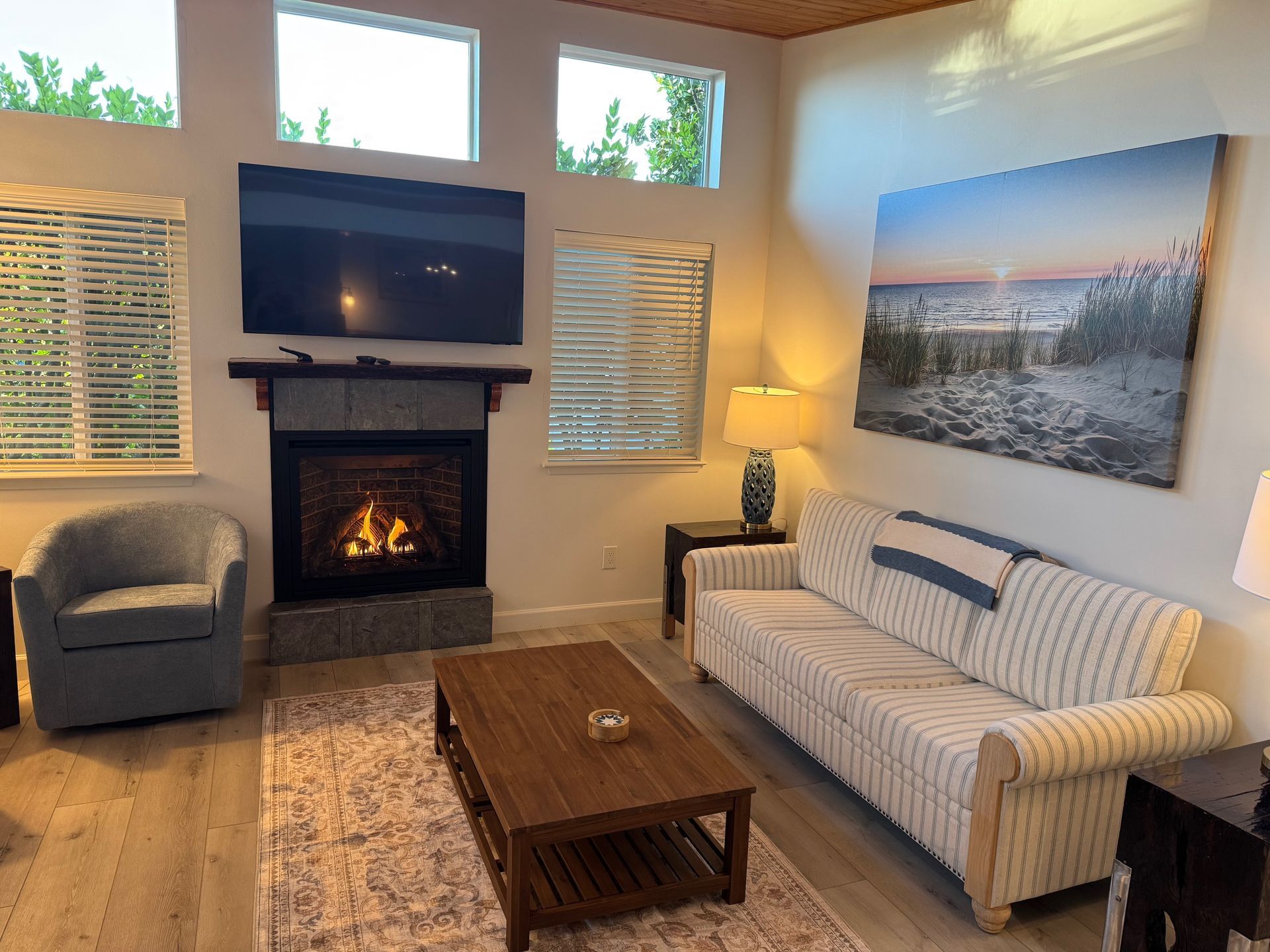 Cozy living room with fireplace, TV, sofa, and artwork. Light wood floors, windows, and neutral colors.