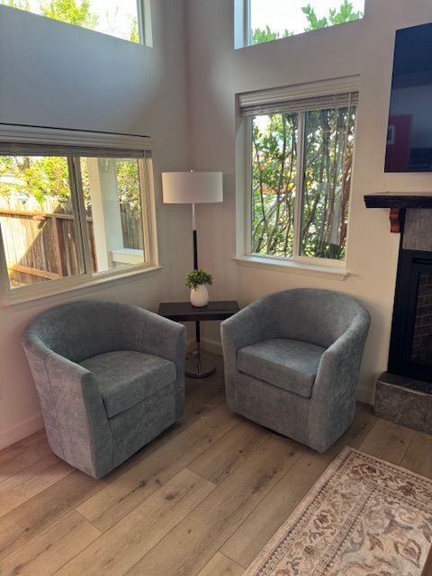 Two gray swivel armchairs face each other, with a table and lamp between them, under windows in a living room.