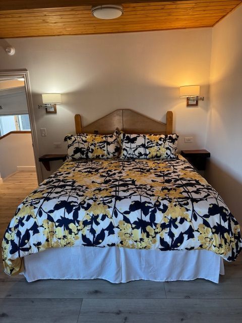 Bedroom with a bed. Floral duvet cover in yellow and navy over a white bedskirt. Two sconces flank the headboard.