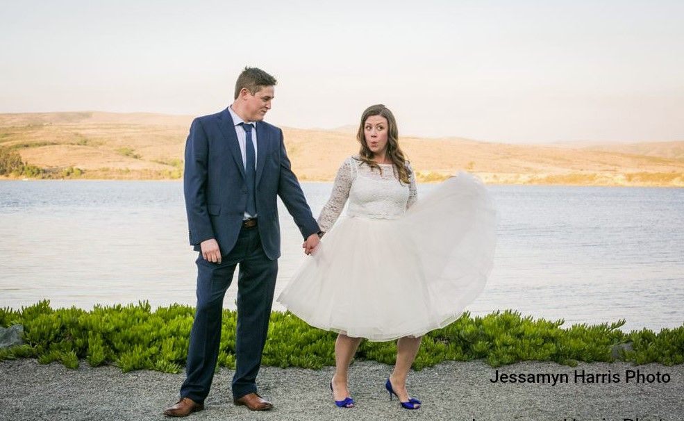 Bride in short wedding dress playfully holds skirt up, making a funny face, while groom looks on by a body of water.