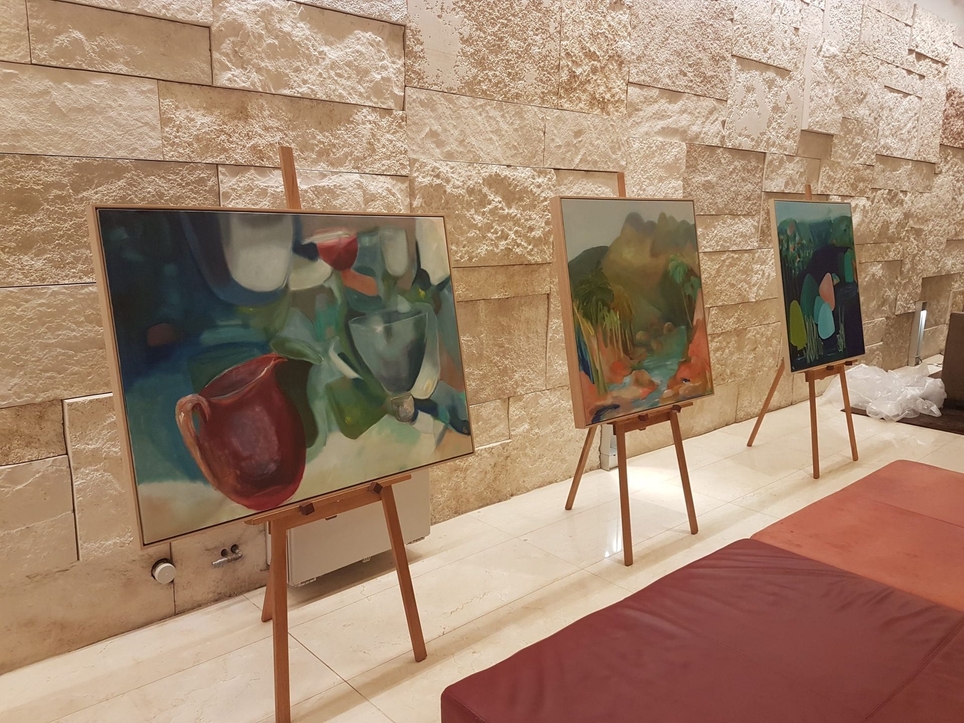 Three paintings are on easels in front of a stone wall.