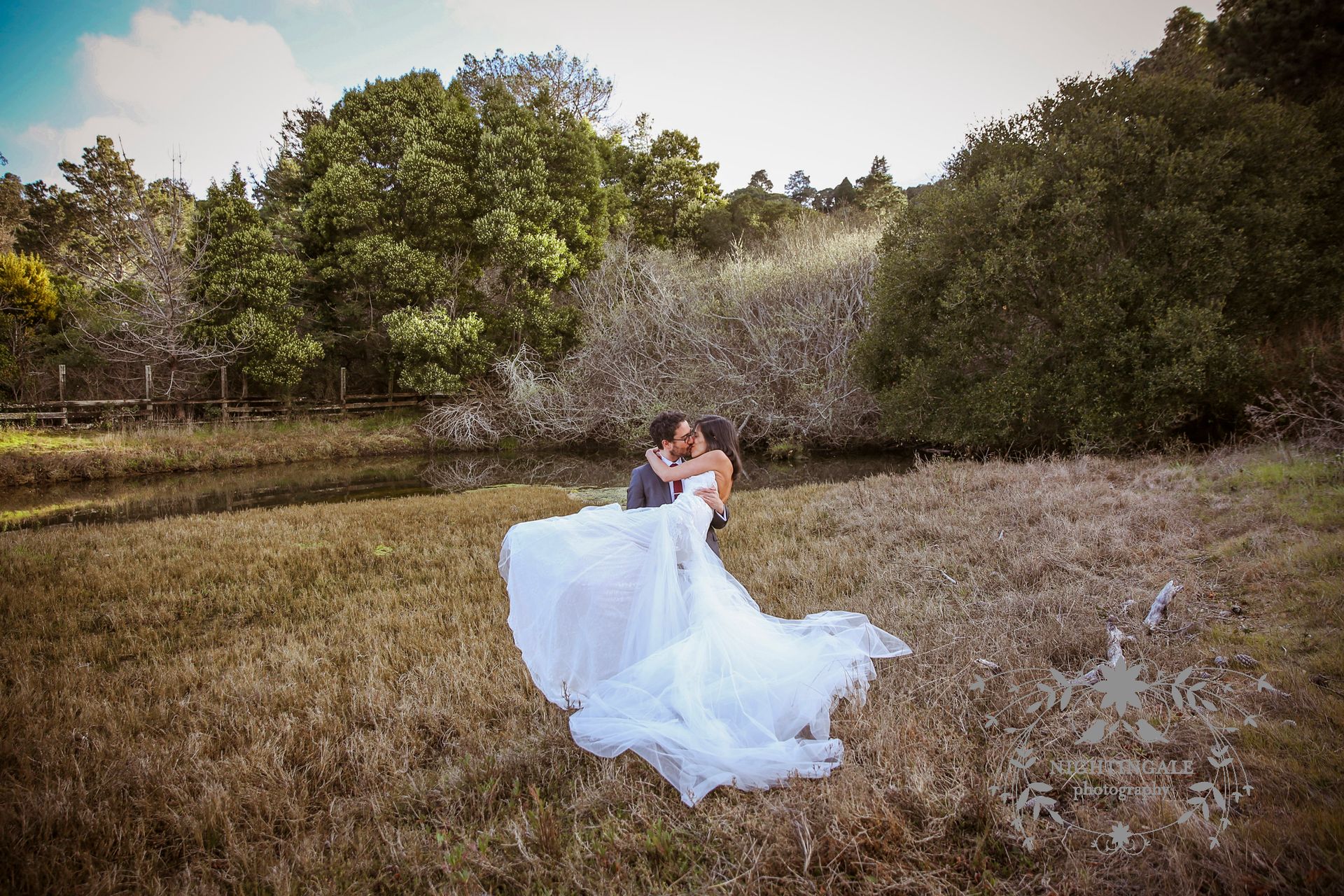 A bride and groom are kissing in a field . the bride is wearing a long white dress.