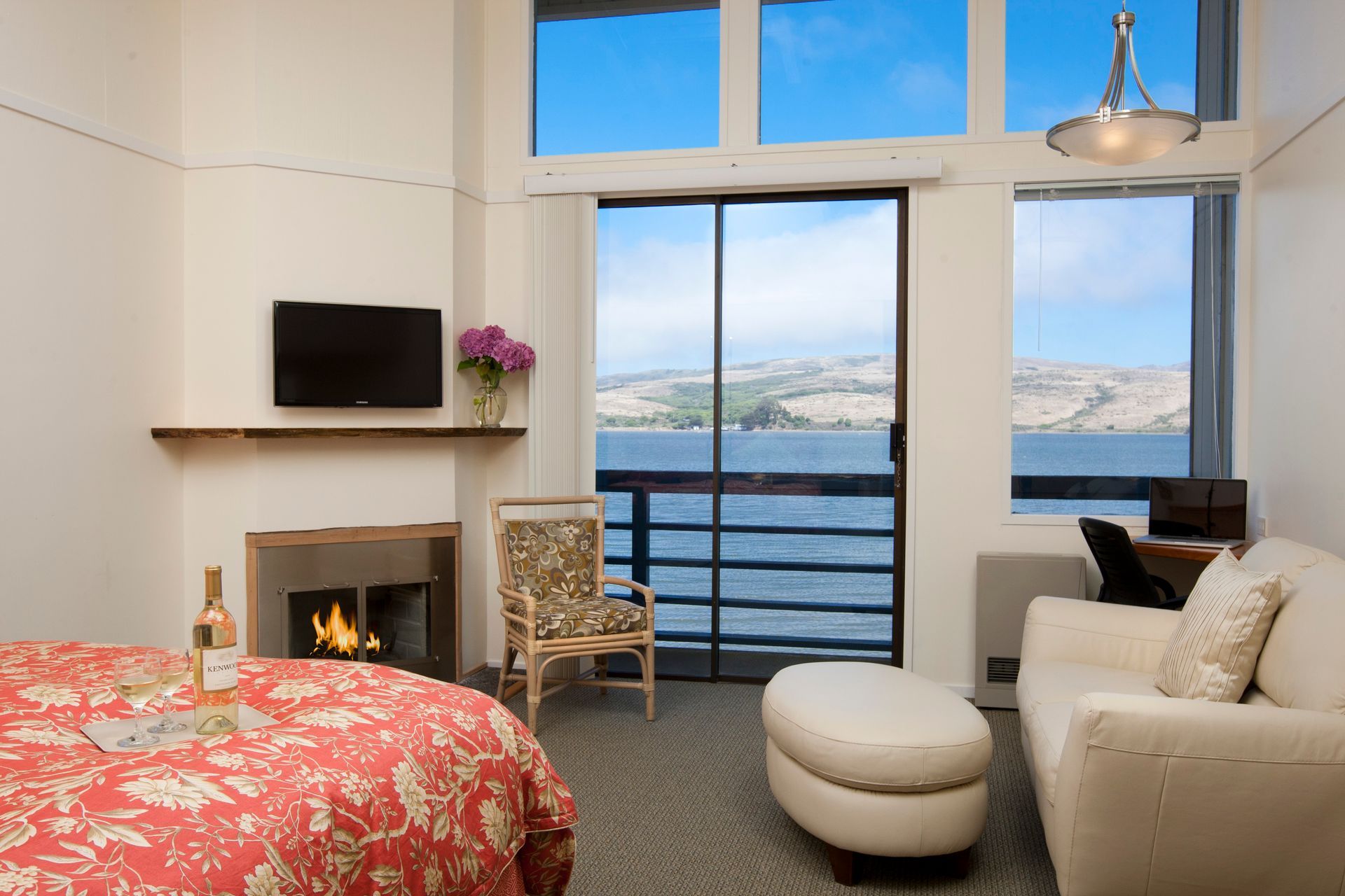 Hotel room with fireplace, water view, sliding glass door, white sofa, and patterned bedspread.
