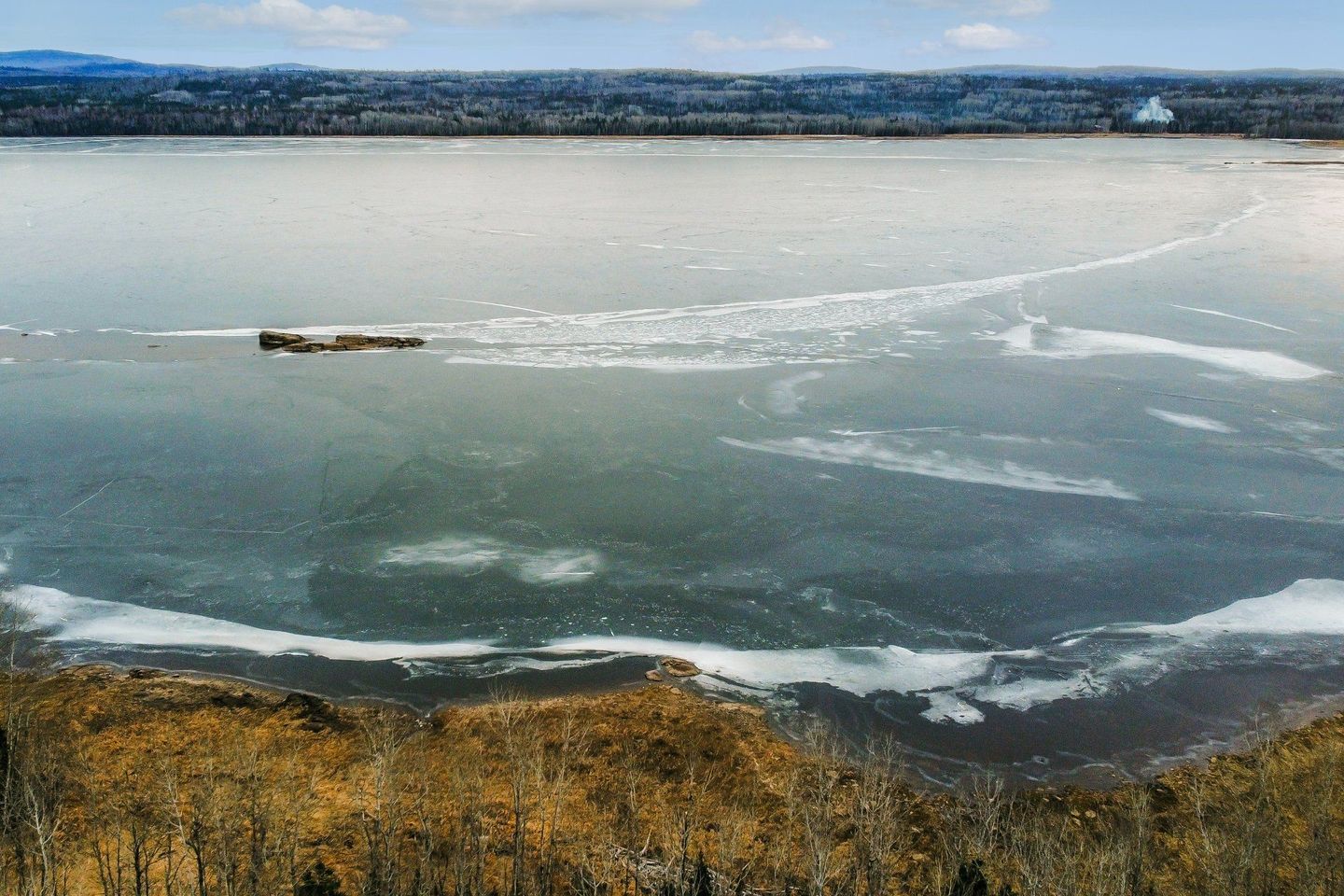 Vue aérienne d'un lac gelé avec des arbres au premier plan et des montagnes en arrière-plan.