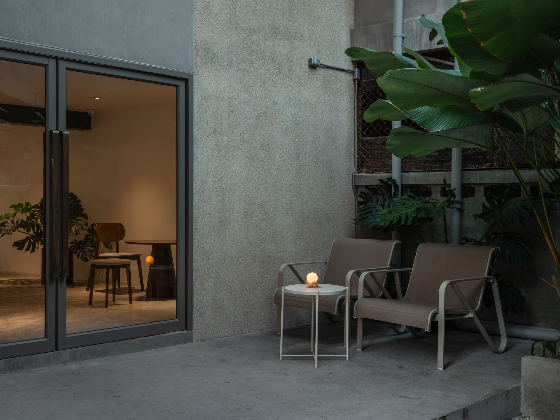 Modern patio with two lounge chairs, small table and candle beside a glass door to a warmly lit interior.