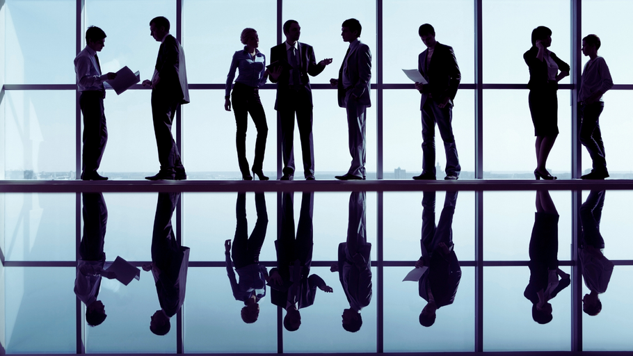 Silhouetted business people standing and discussing in a modern office with reflective flooring and large windows.