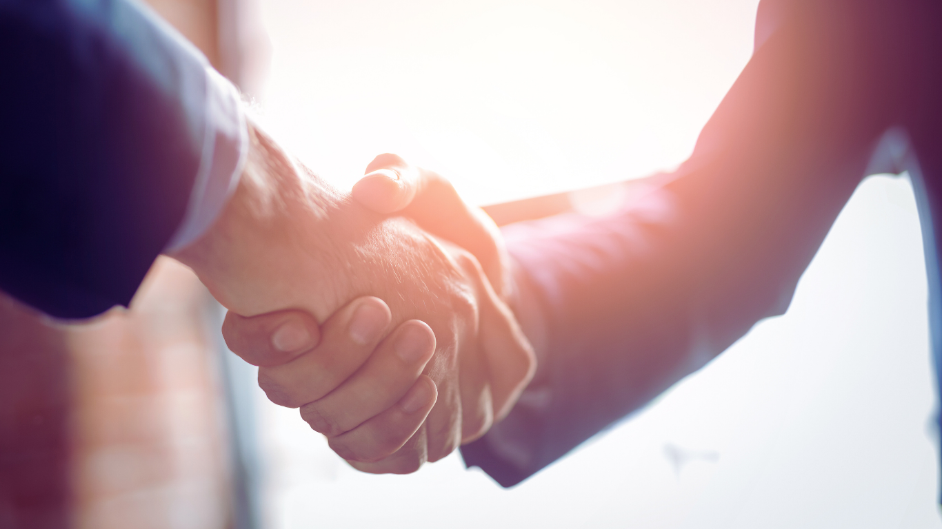 Two people in suits shaking hands, backlit by sunlight.