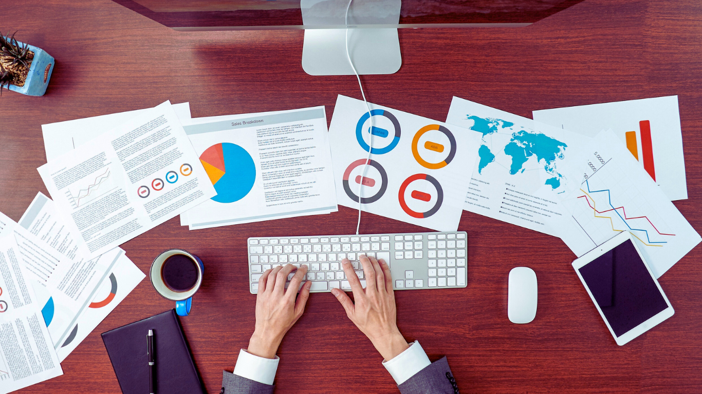 Person typing on a keyboard at a desk covered with financial charts, a tablet, and a coffee cup.