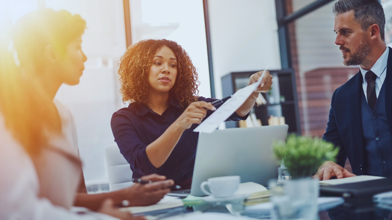 Business meeting: woman pointing at document, laptop, two colleagues, and sunlight.
