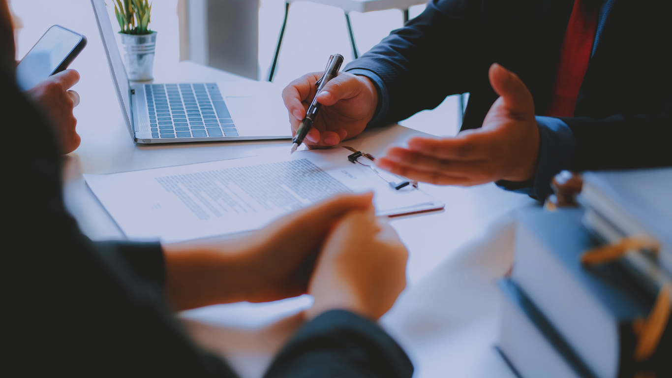 Person signing a document, being assisted by someone else, with a laptop and plant on a desk.