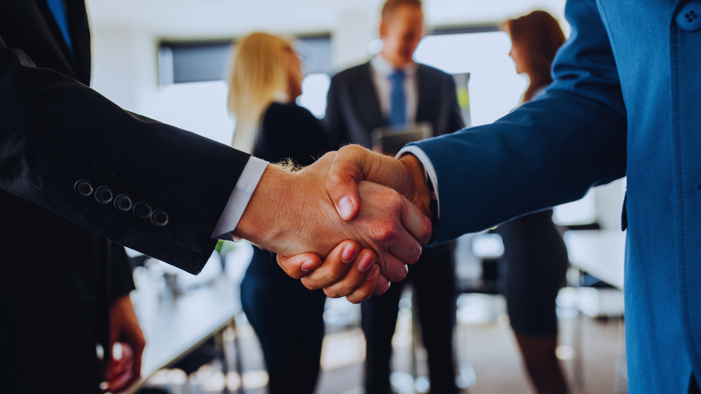 Two businesspeople in suits shaking hands, office setting, colleagues in the background.