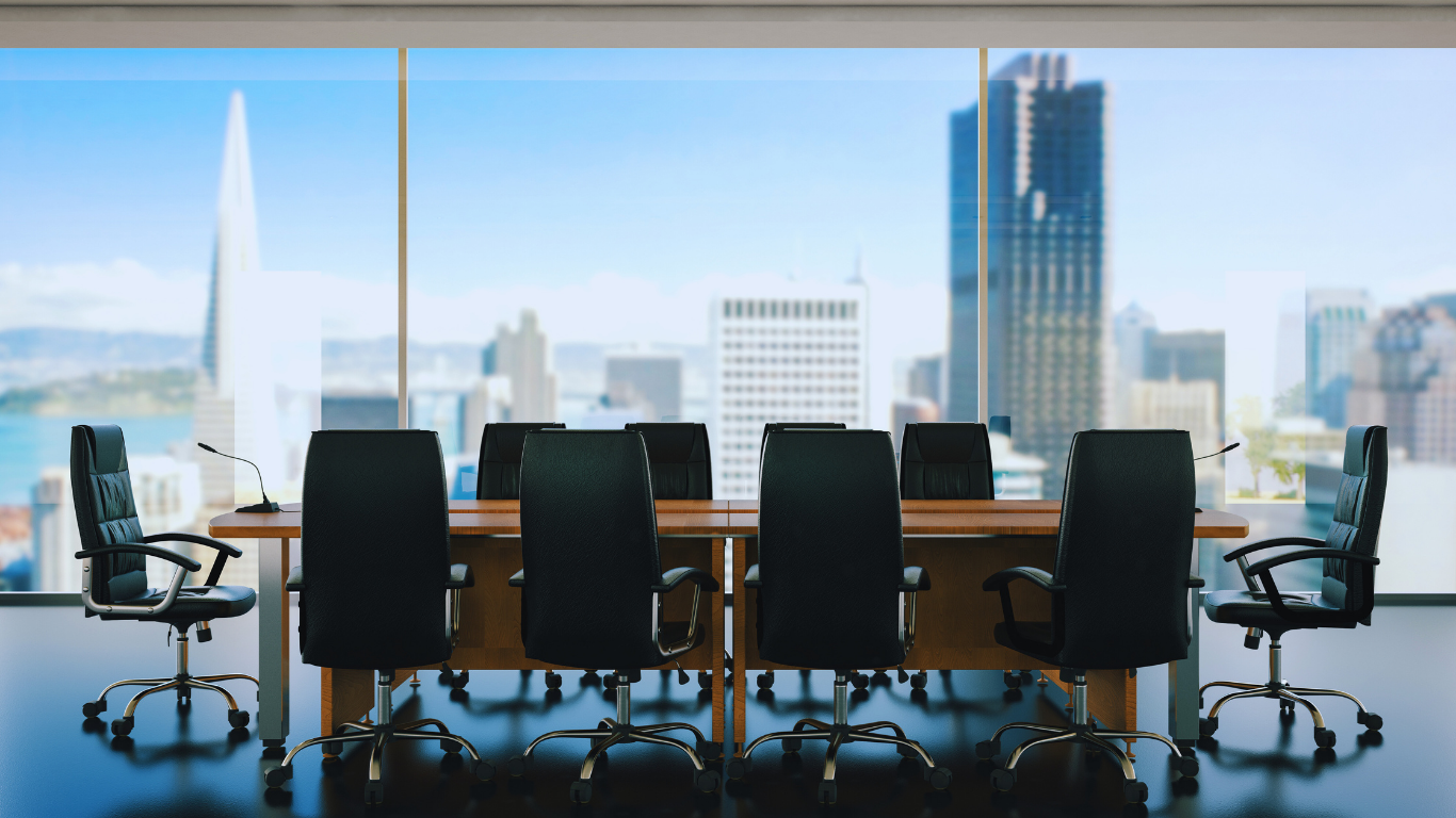 Empty boardroom with black chairs around a wooden table, large windows show a city skyline.