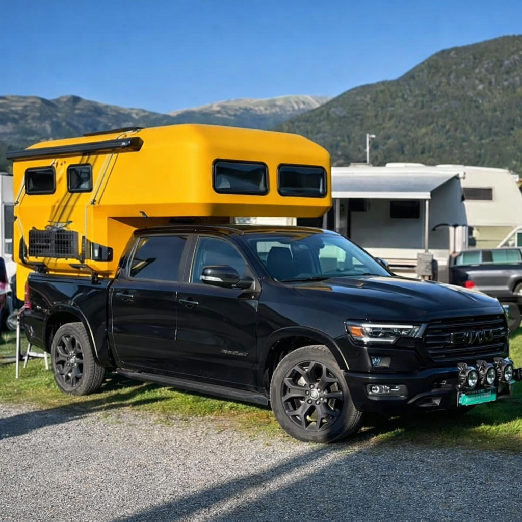 A black pickup truck with a large, bright yellow camper unit mounted in the truck bed, parked in a gravel lot.