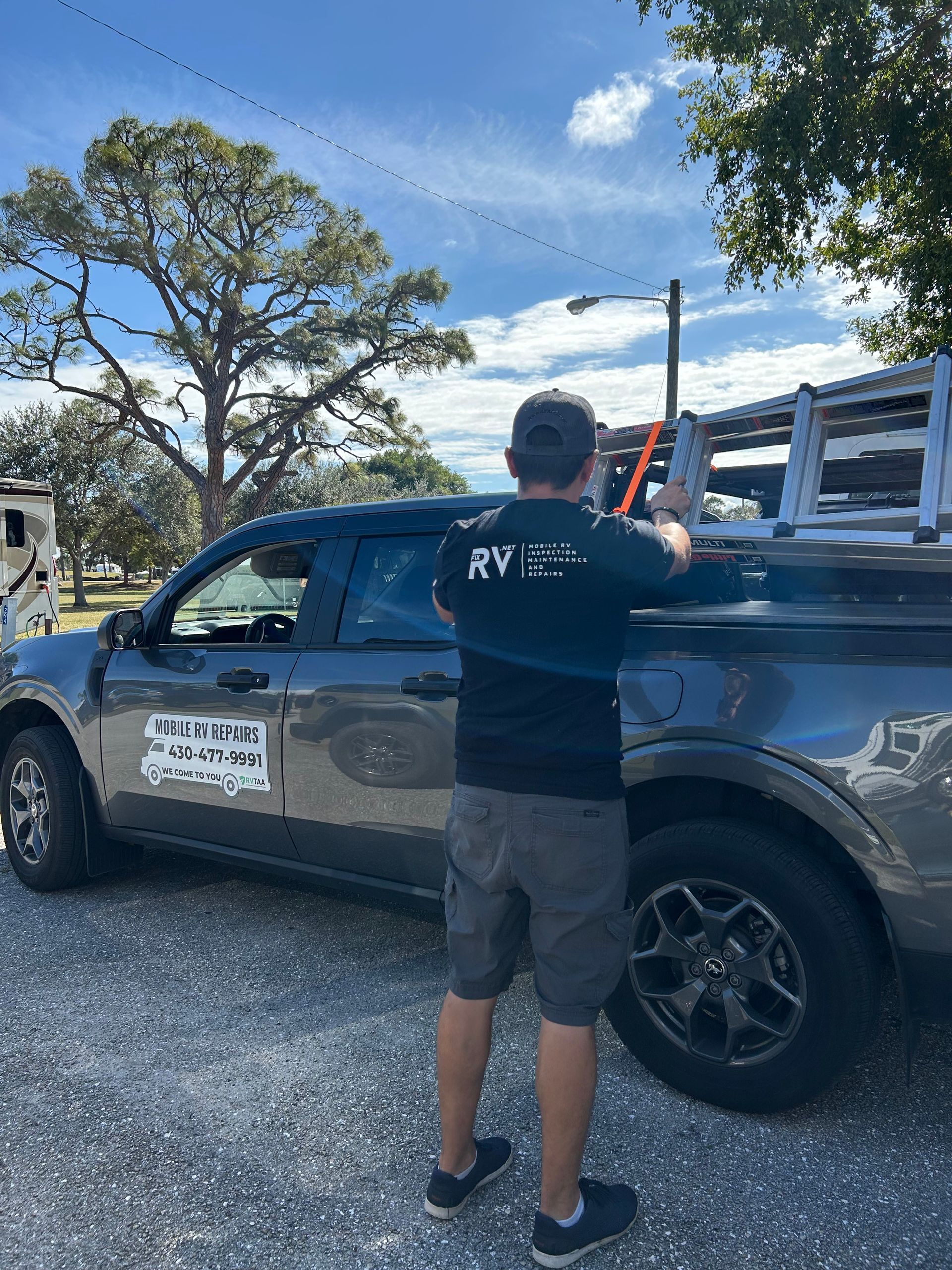 A person in a black shirt and shorts secures a ladder to the roof rack of a gray pickup truck on a gravel lot.